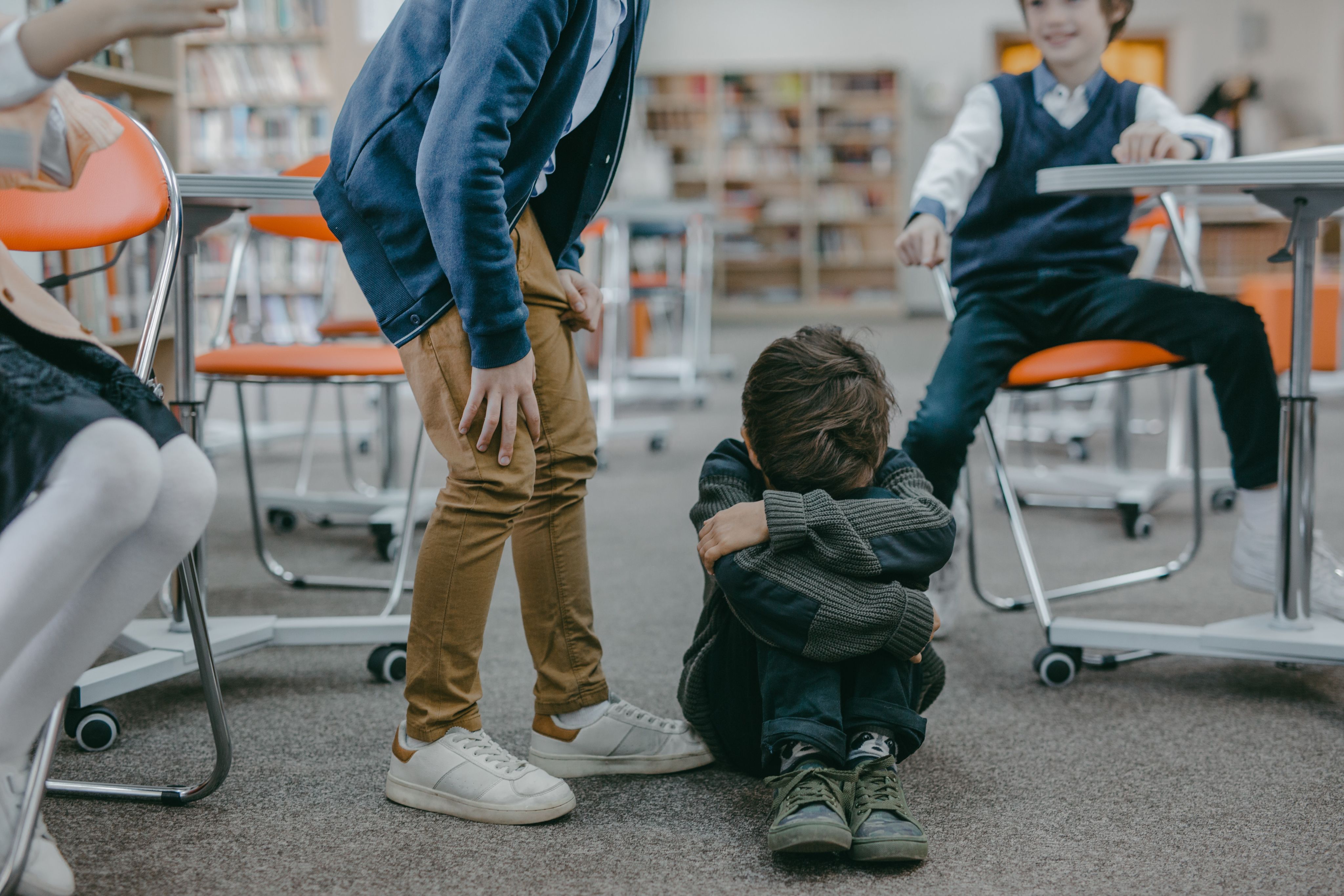 Young child crouched with their head in their knees while another child is standing over them, another pointing at the child crouched. 