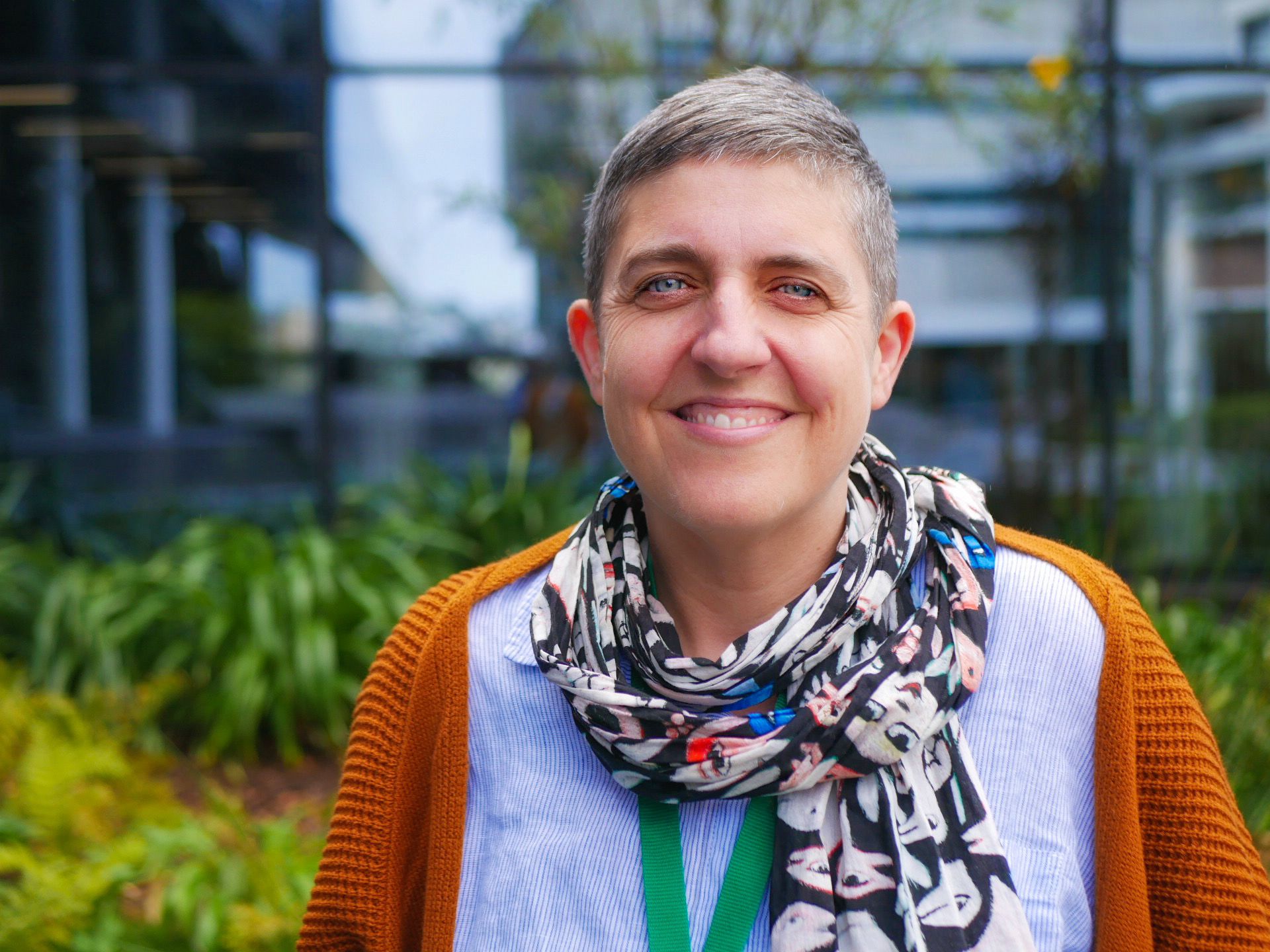 Person with colourful scarf, very short grey hair, and a orange cardi