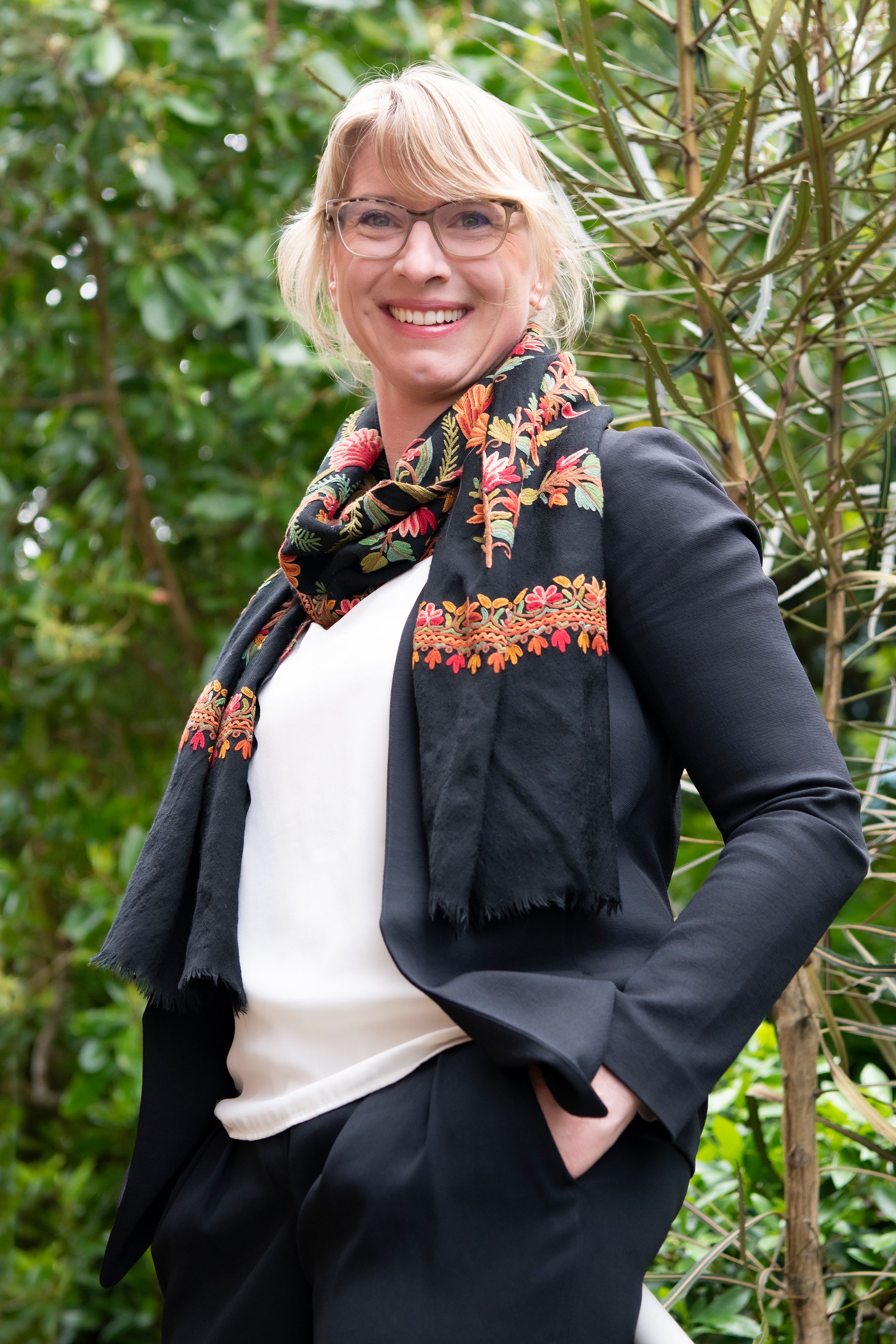 Person with blonde hair in ponytail and glasses, wearing bright scarf