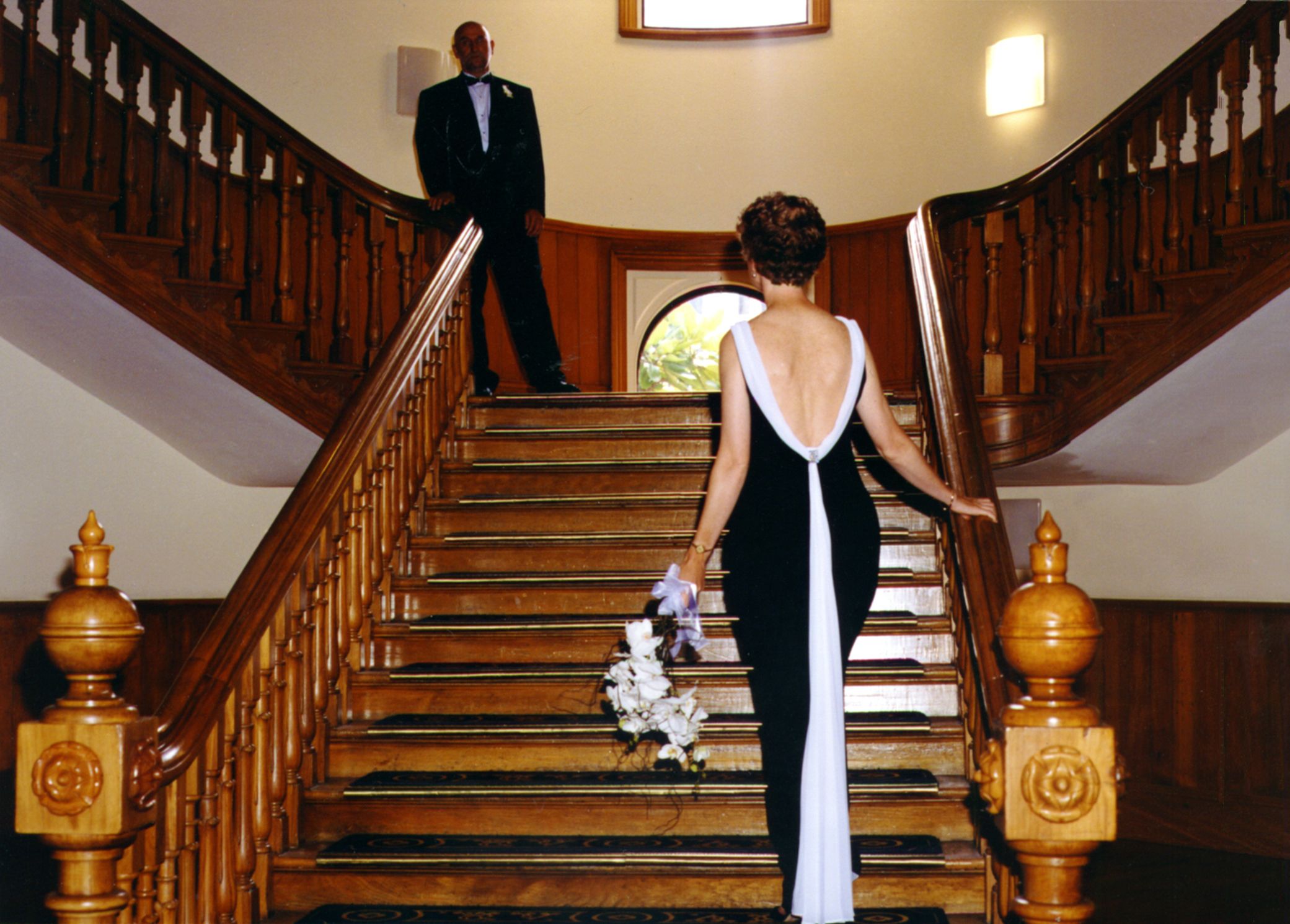 IMage of woman in black and white wedding dress holding floral arrangement looking up stairs to man in black suit