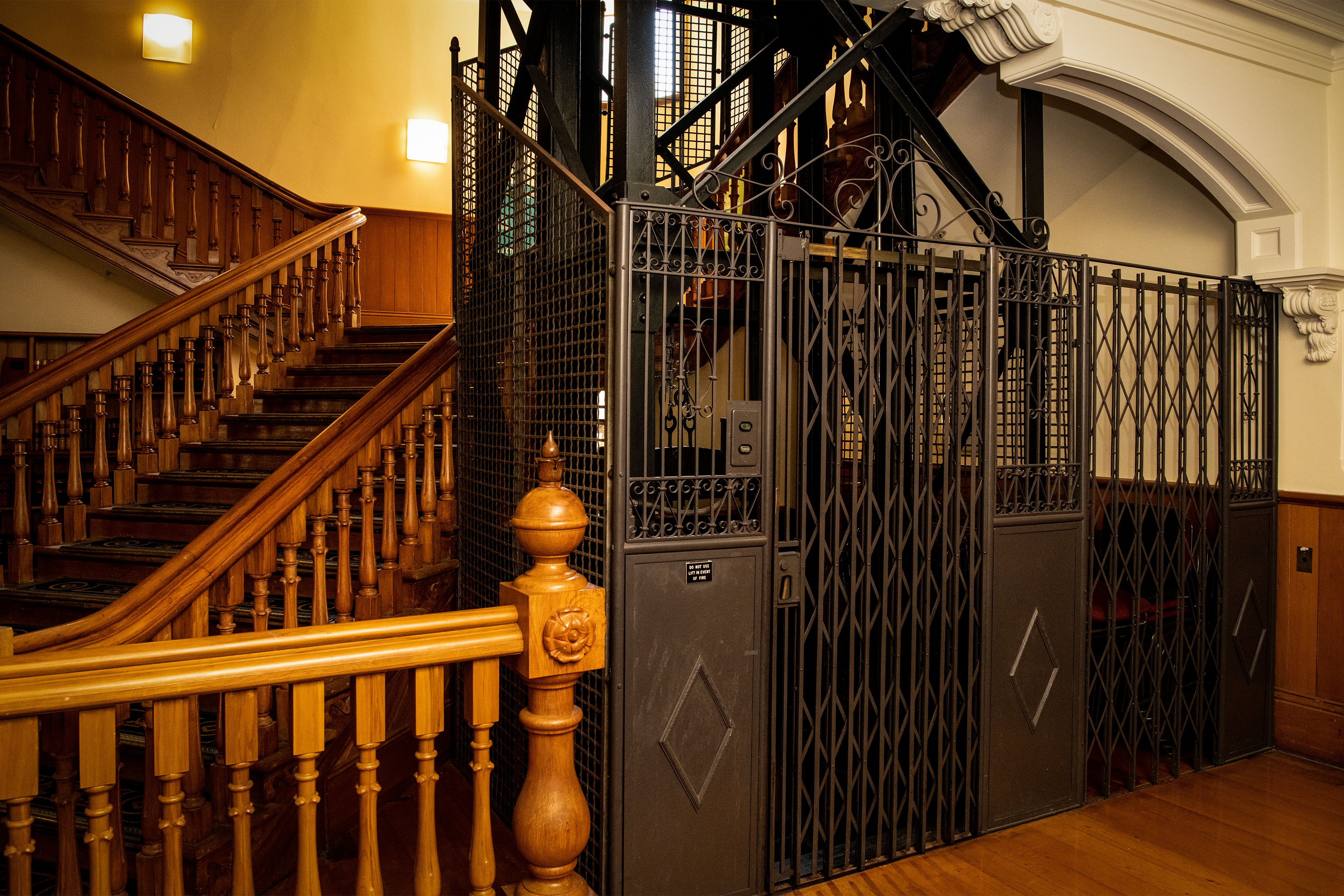 Black-painted metal lift with a decorated metal frontage framed against wooden stairs