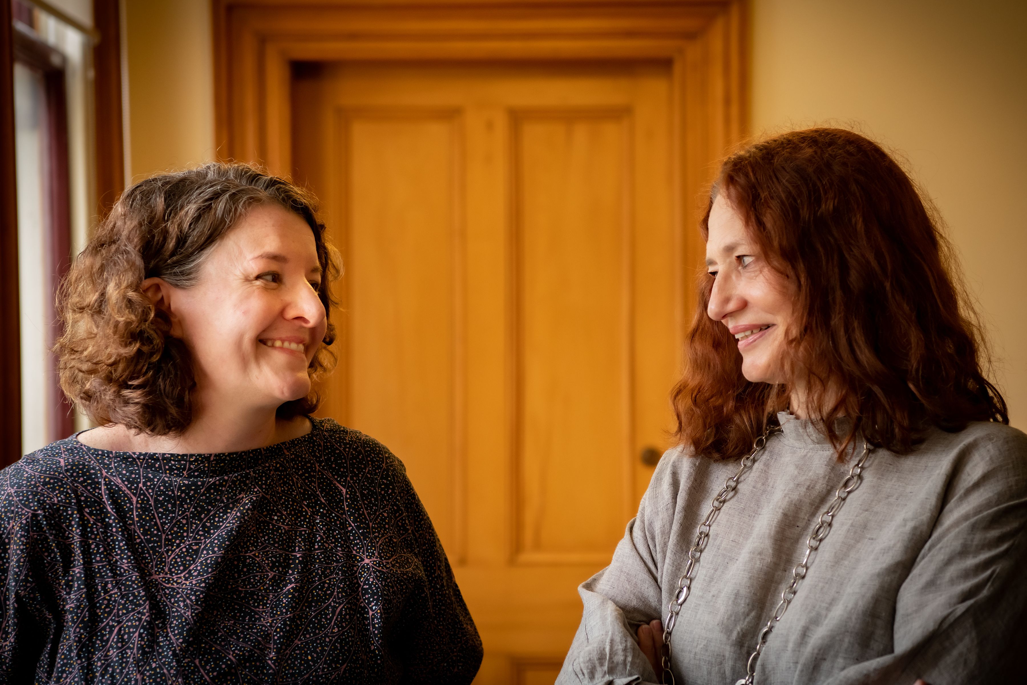 Two curly-haired women, one smiling at camera wearing navy and pink top, the other smiling at the one of the left, wearing grey linen top with long silver necklace
