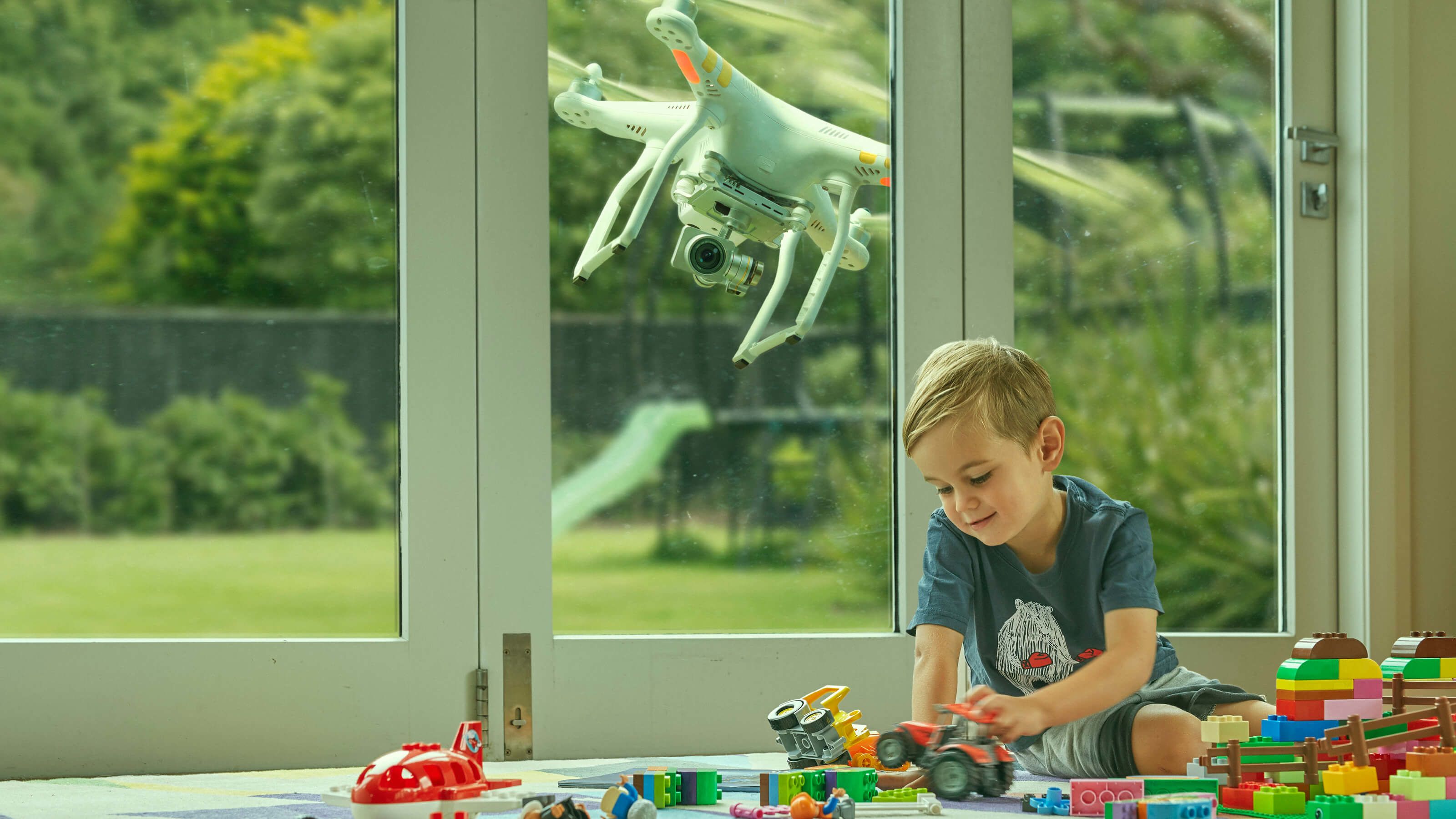 A small boy playing with his toys in front of a sliding doors and a drone is hovering outside of the doors watching his every move.