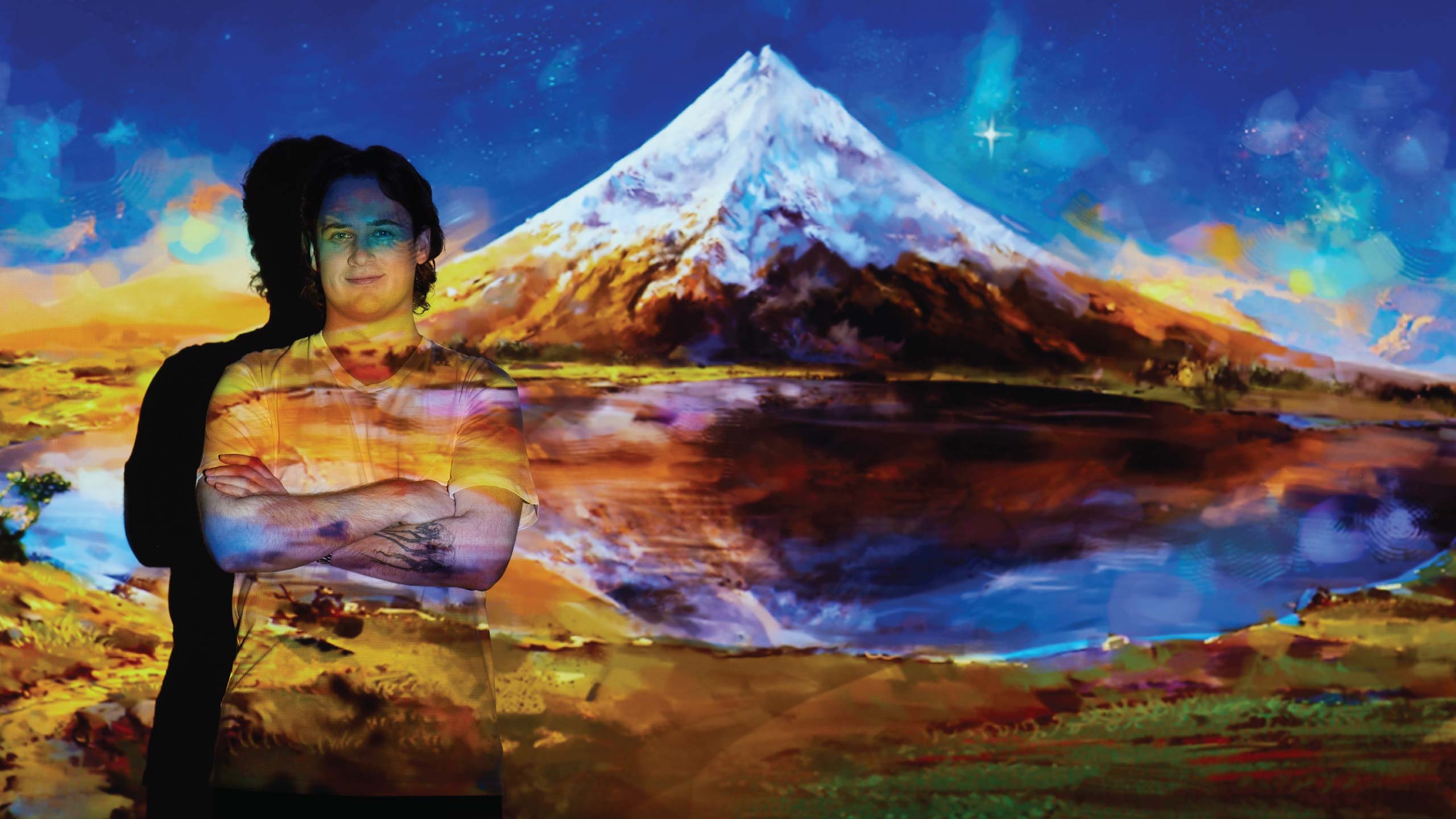 Tom Trengrove standing with arms folded looking to camera with one of his digital landscape paintings is projected onto him