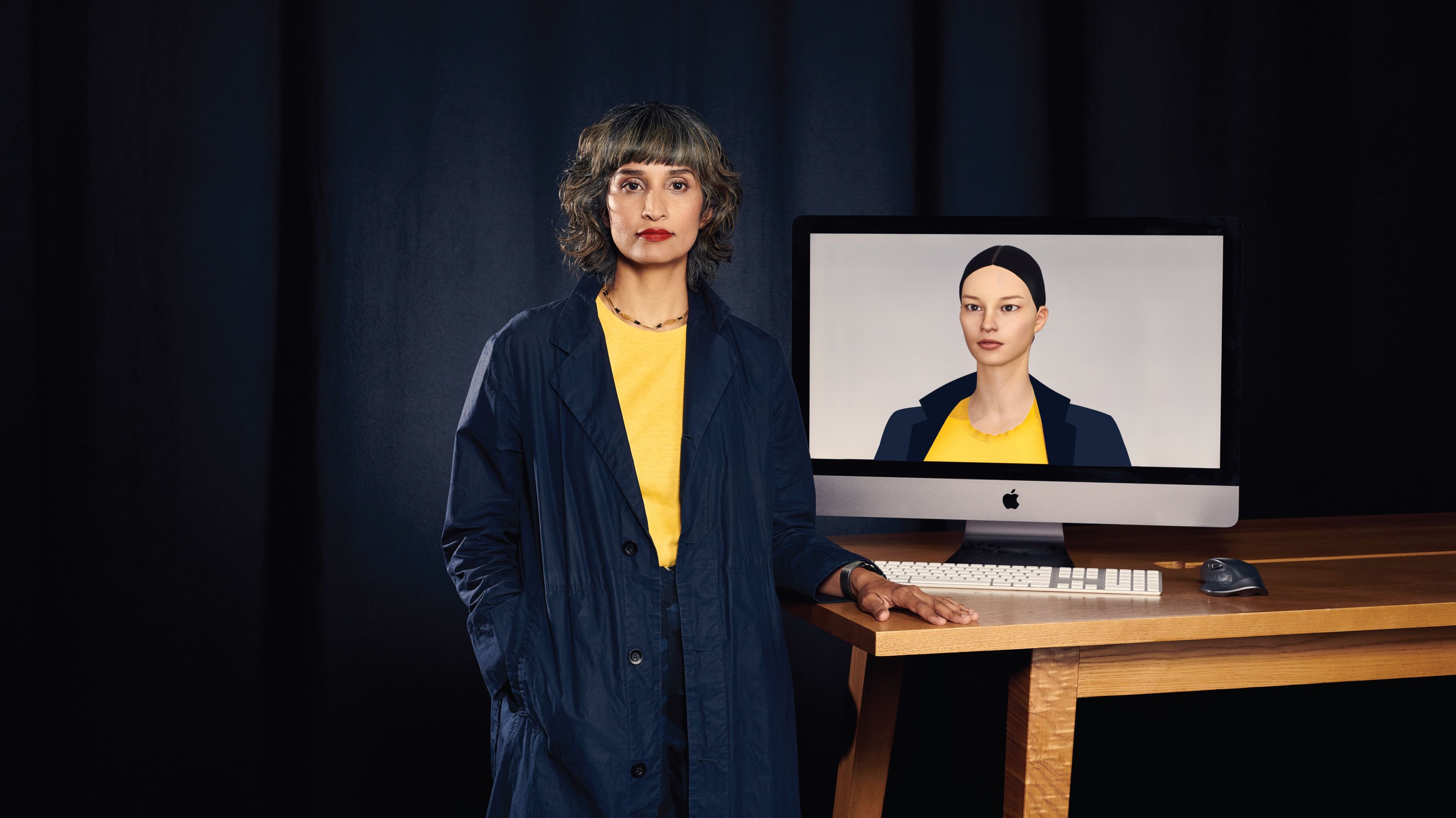 Raqi Syed stood in black room next to iMac showing her white digital human avatar